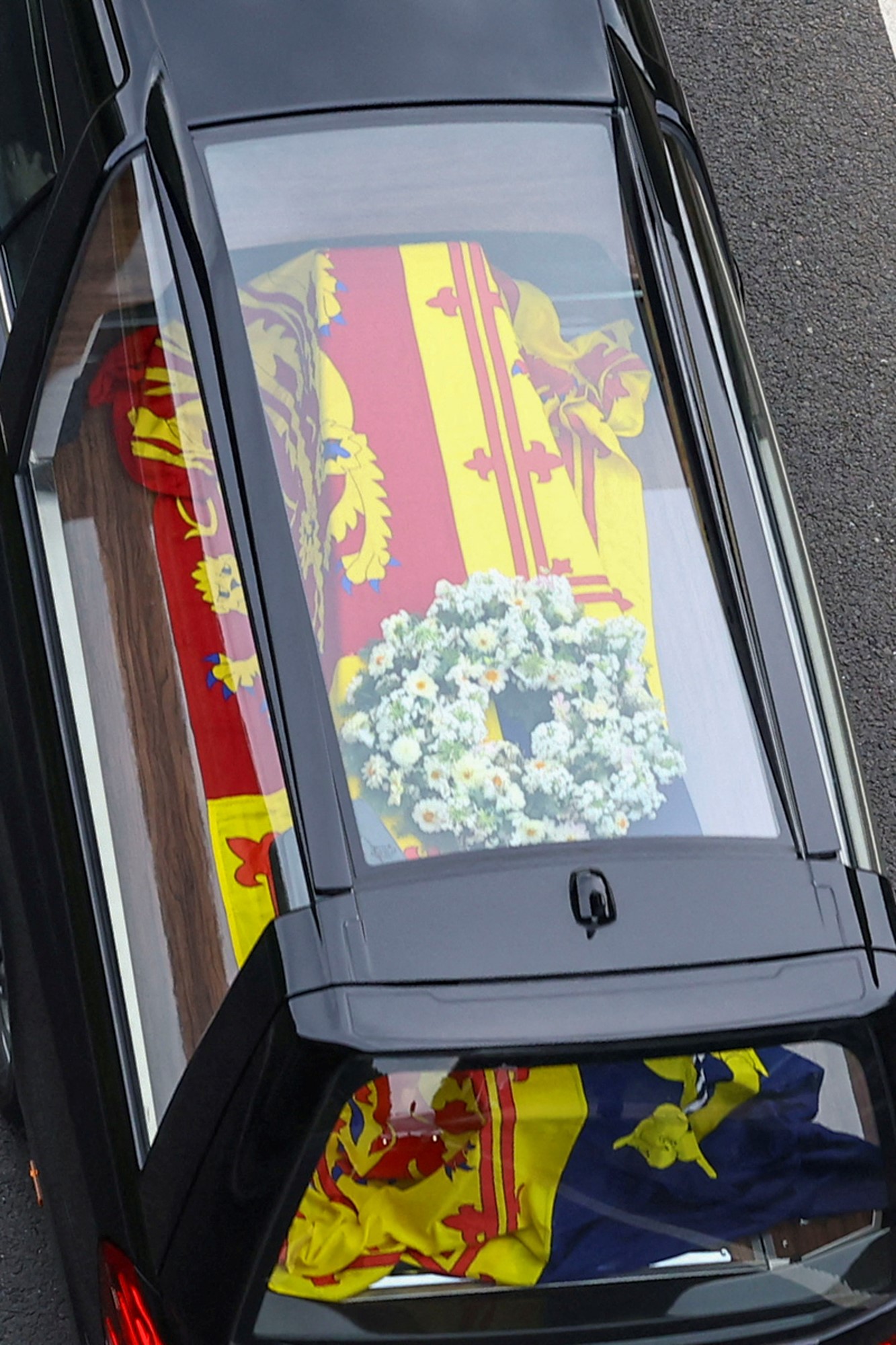 A picture of the Queen's coffin from above, seen through the glass roof of the hearse carrying it. It is lying underneath a brightly coloured flag.