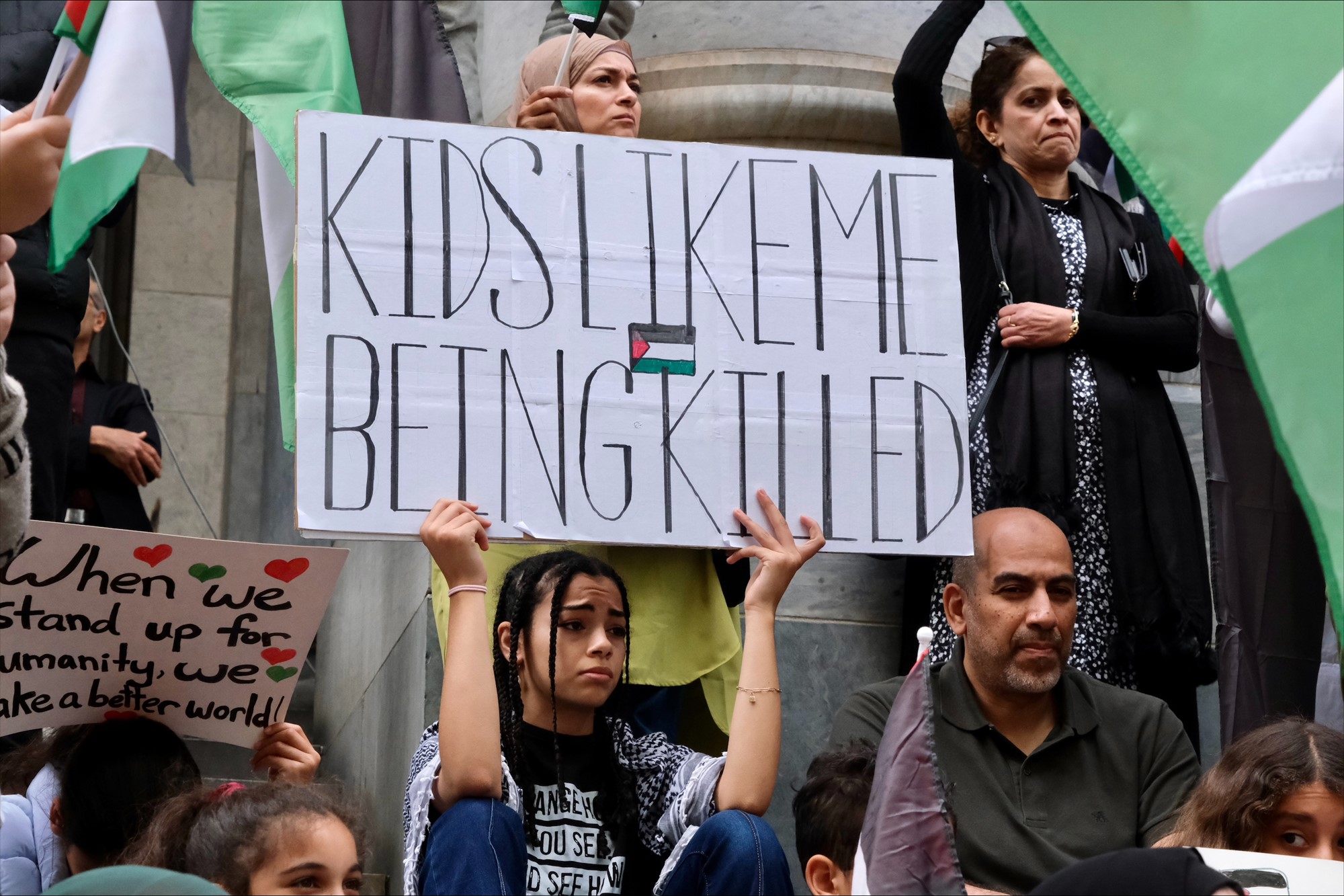 A girl with braids holds a sign saying "Kids like me being killed".