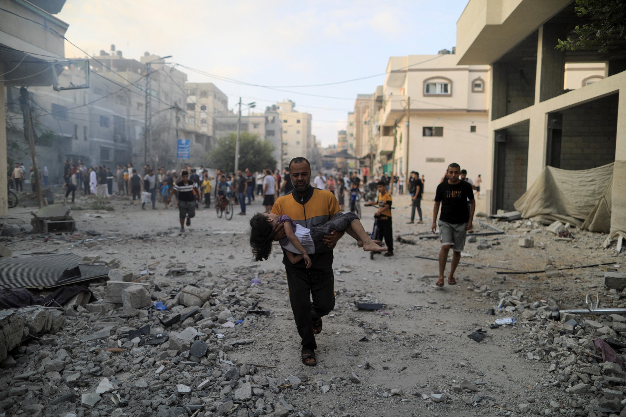A man carries a child in his arms, down a street strewn with rubble