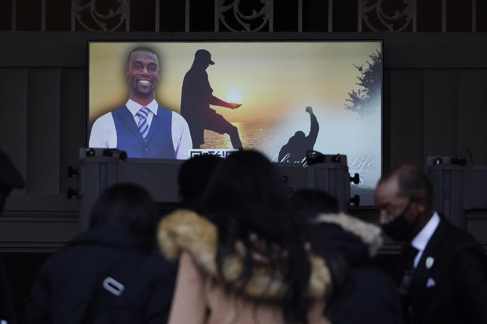 People file into a hall with a screen showing photos of a black man.