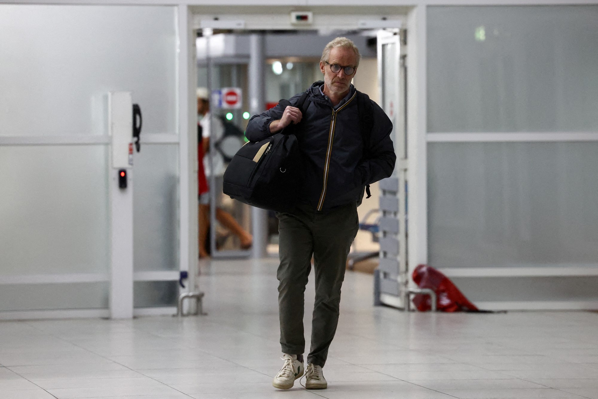 A man with grey hair, glasses, and a bag walks thorugh the arrivals gate