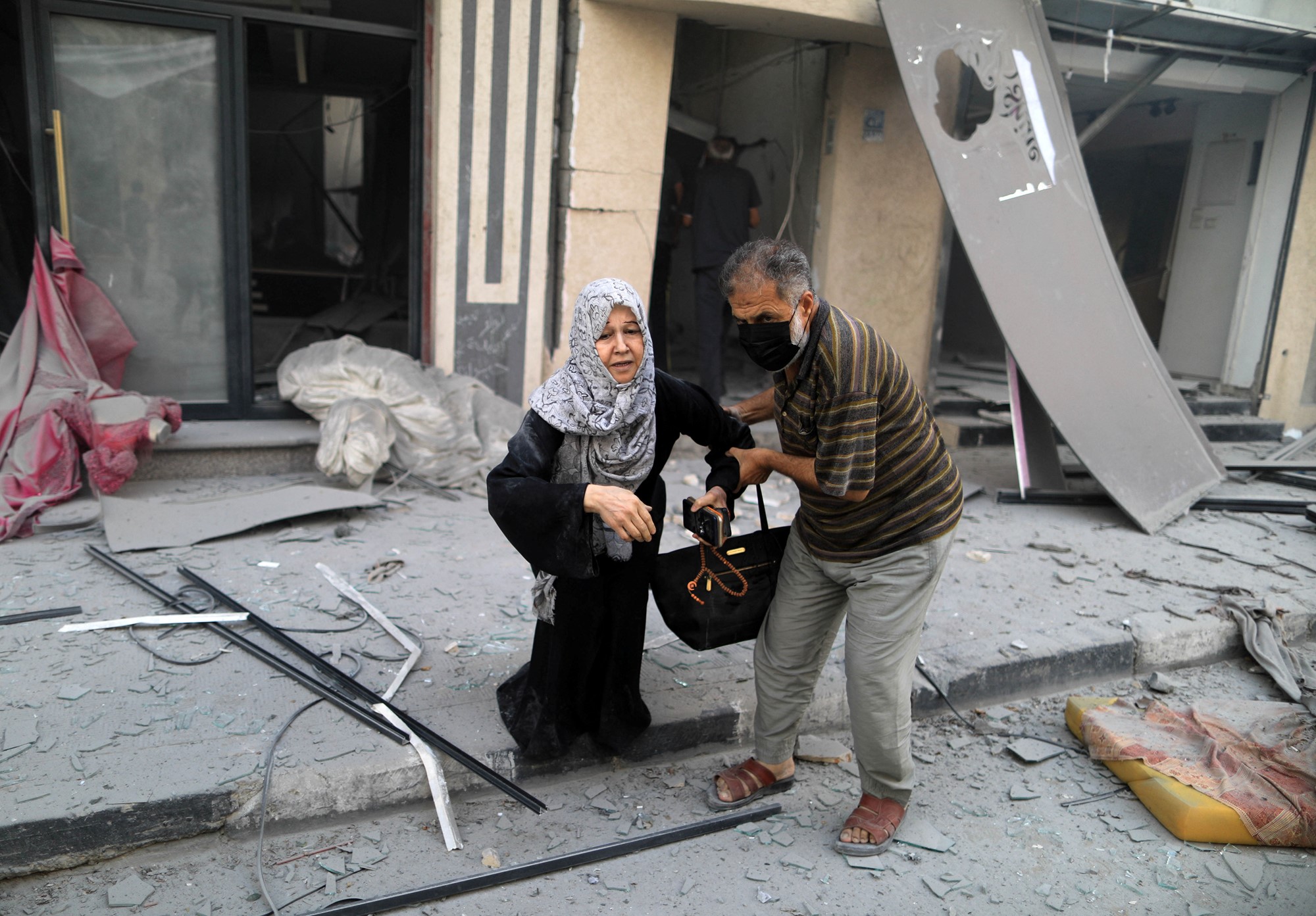An older man helps hold up an older woman as she stumbles, outside a destroyed building