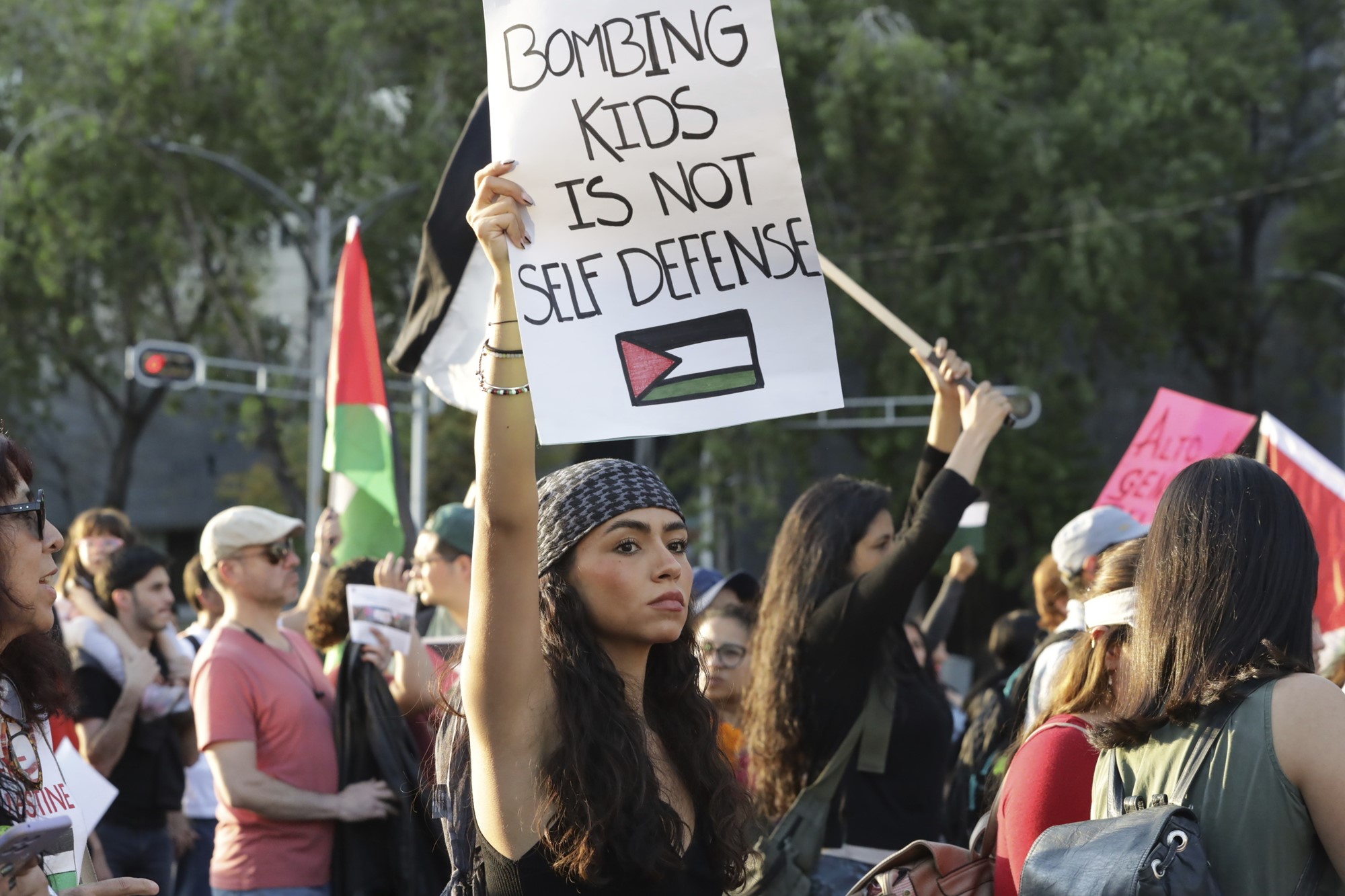A young woman holds a sign that reads "bombing kids is not self defense" with a Palestinian flag drawn underneath