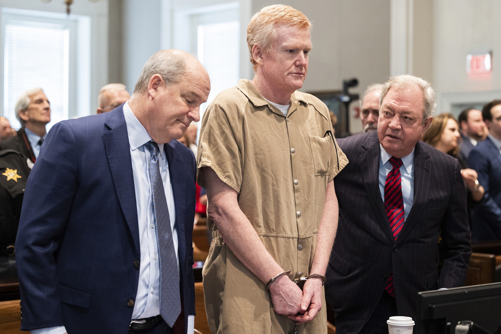 A man with blond hair is wearing a beige jumpsuit and shackles. Next to him are men in suits.