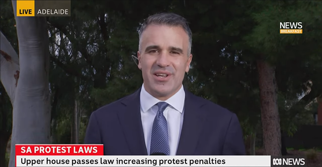 A middle-aged man in a suit doing a TV interview outdoors near trees