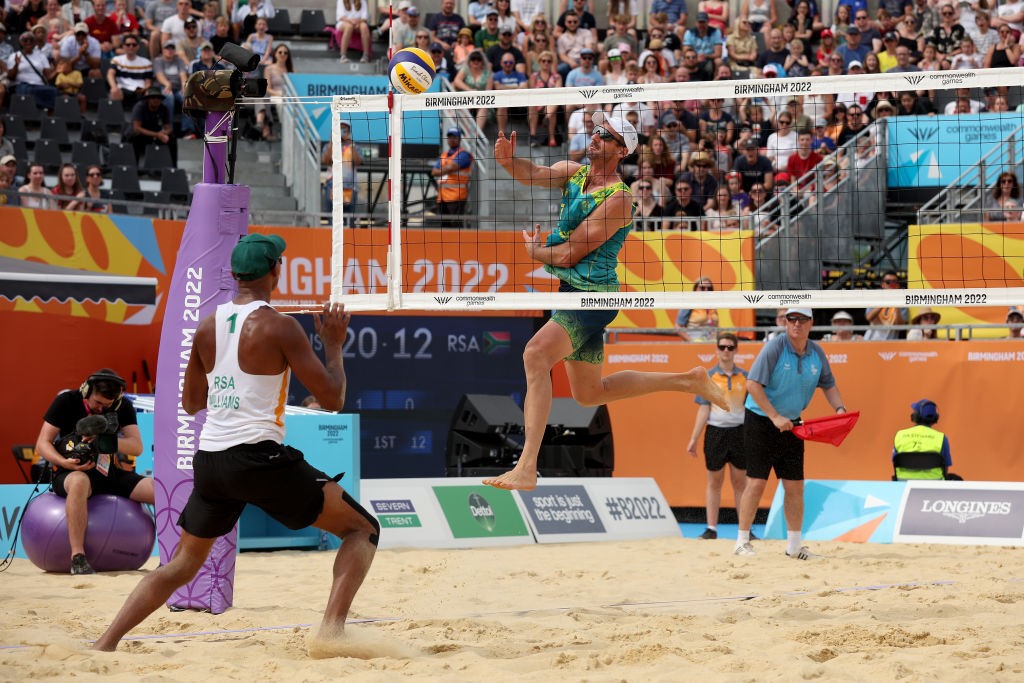 australian beach volleyballer chris mchugh jumps into the air gesturing towards a volleyball, another player prepares to receive the ball on the other side of the net