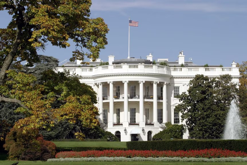 A large, white stone colonial building surrounded by greenery and flying an American flag.