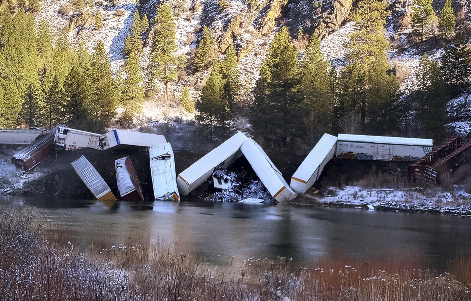 A photo of train cars which have fallen from a track and into water below. Snow and trees on a steep hill in the background