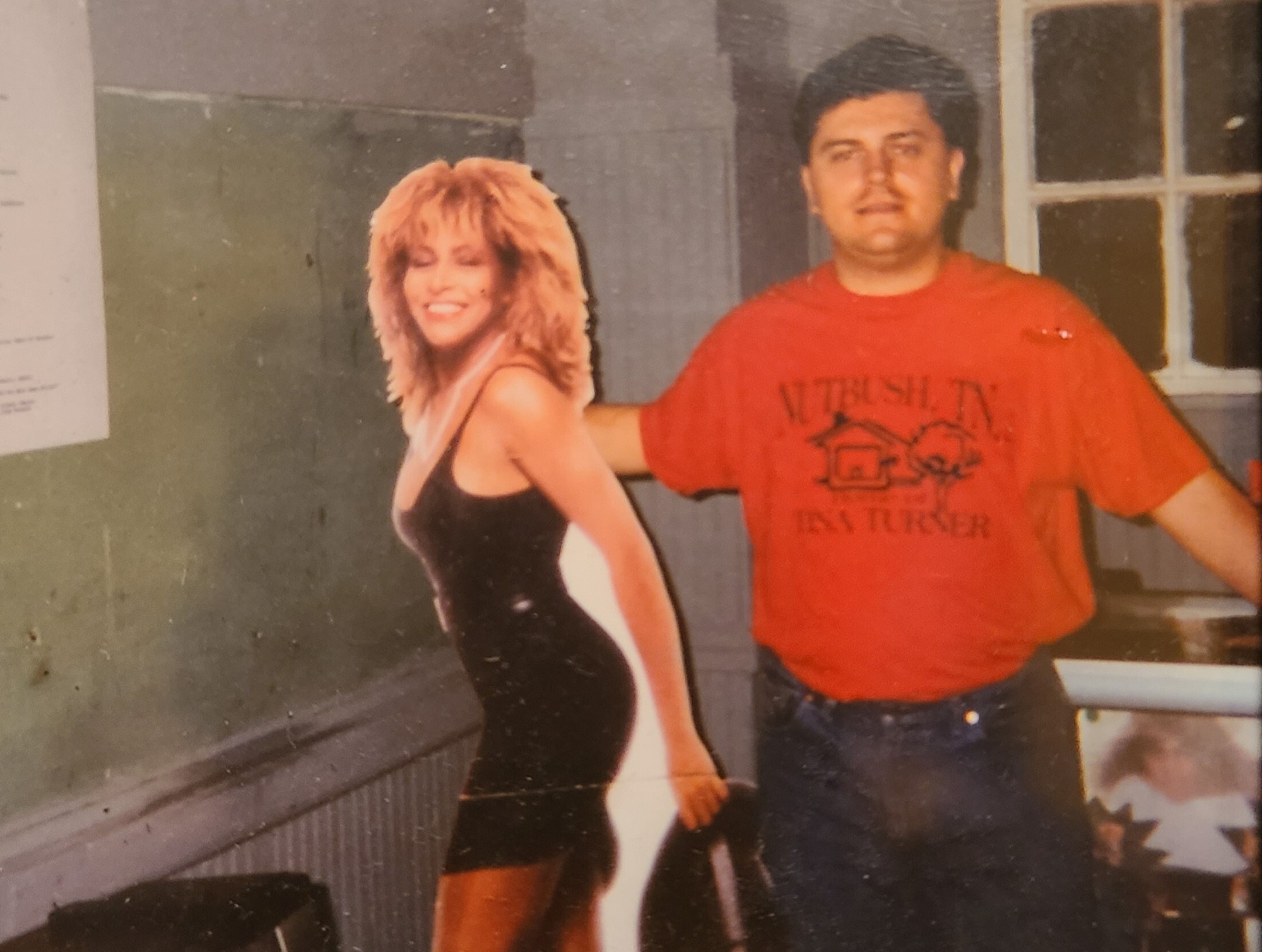 A man wearing a red tshirt poses next to a cardboard cutout of Tina Turner.