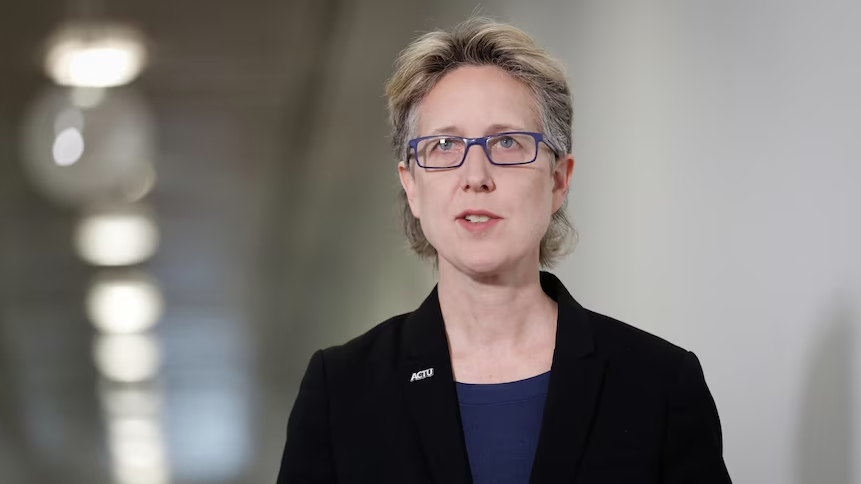 A middle-aged white woman with glasses and short hair speaks in a white hallway.