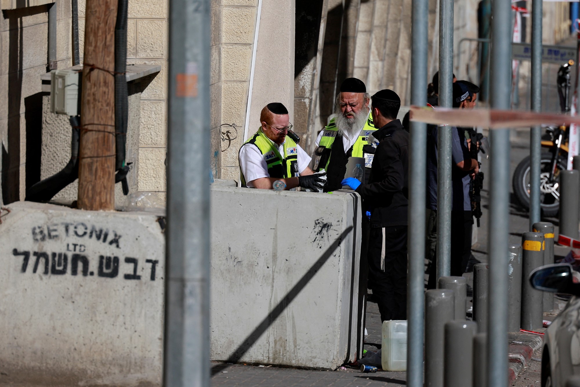 Two older men in hi-vis vests and yarmulkes stand behind a concrete barrier on a city street.