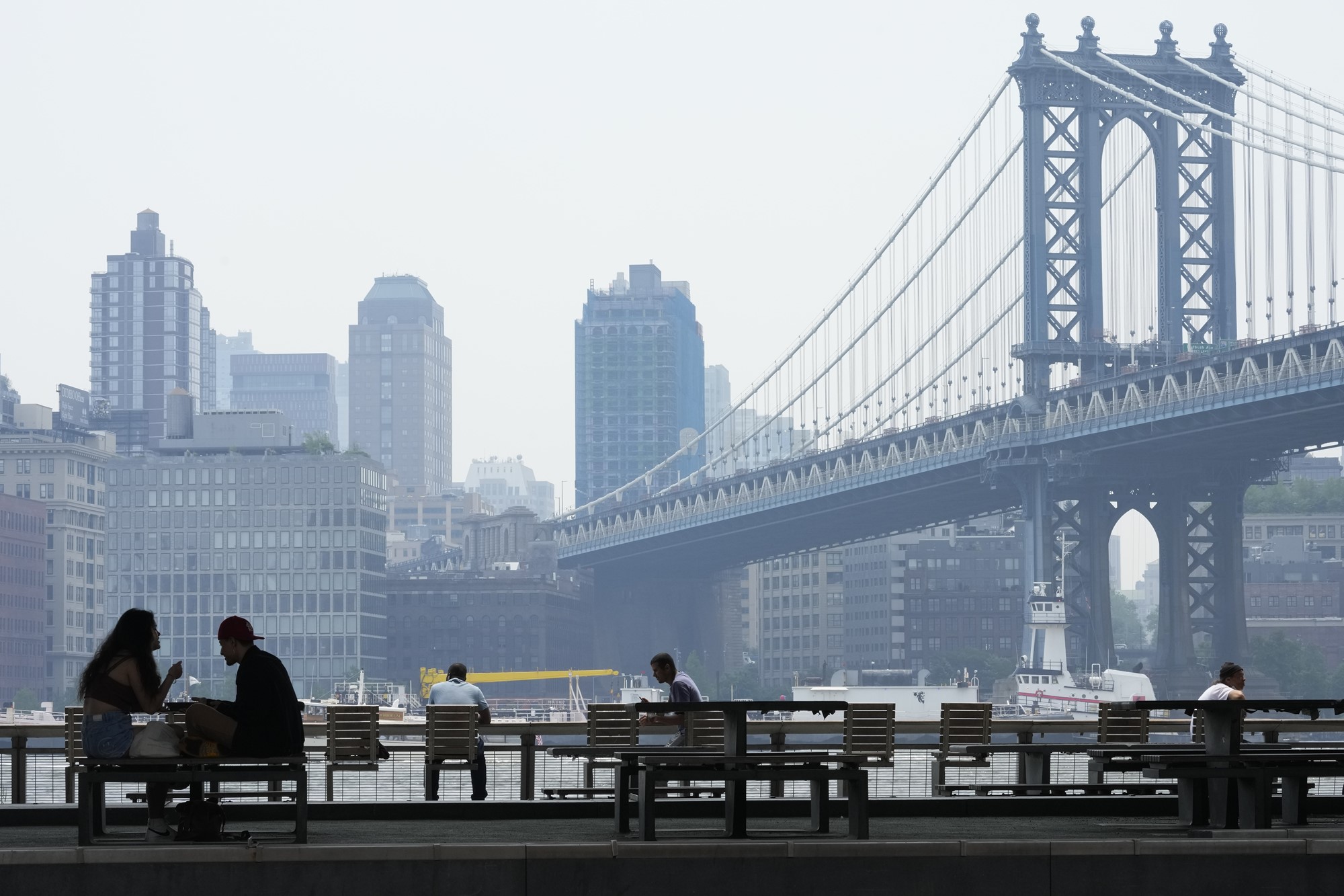 People on the East River promenade are framed by the hazy Brooklyn waterfront skyline and the Manhattan bridge
