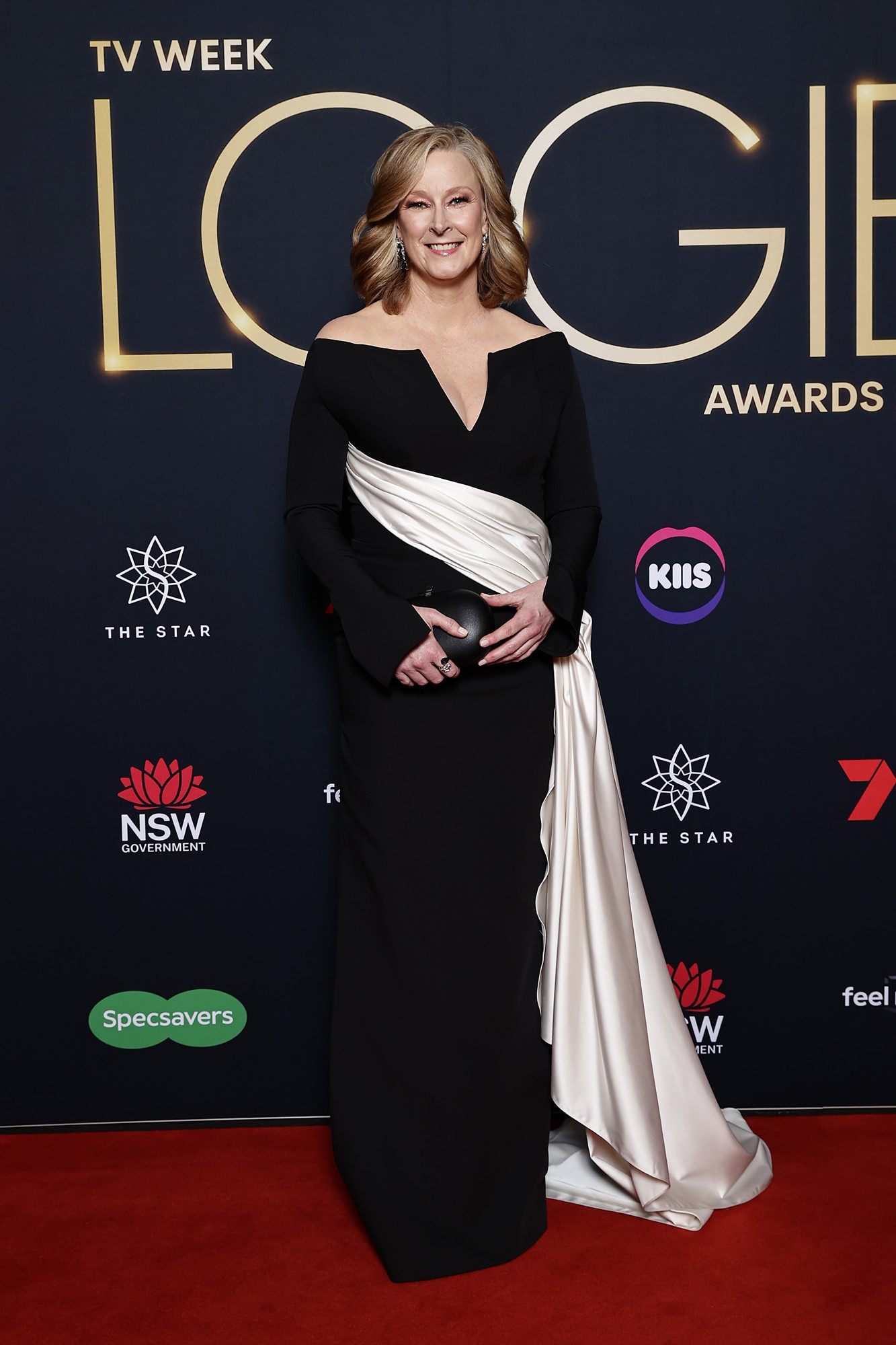 A woman with a long black dress with a white sash, smiliong and posing on the red carpet