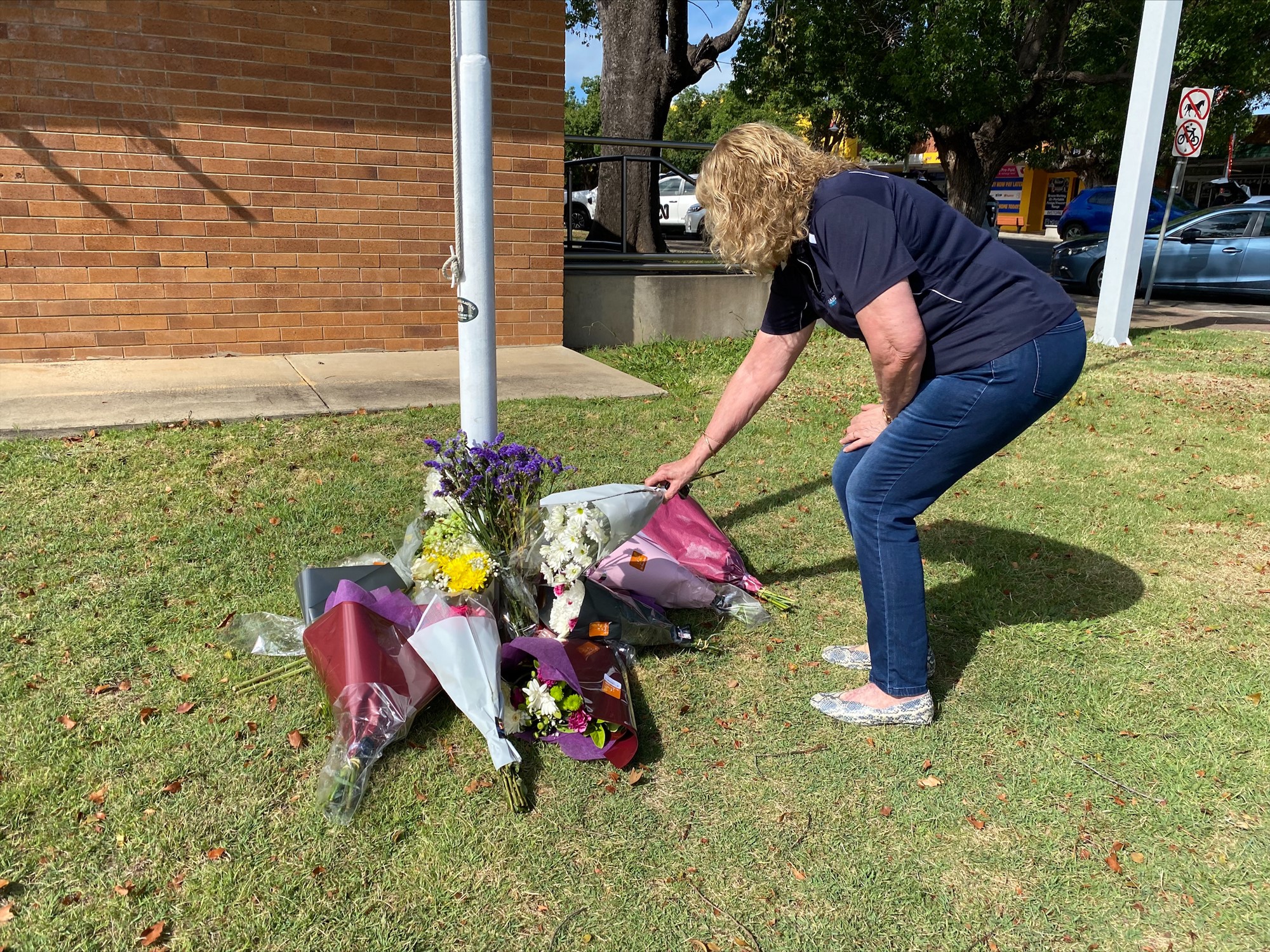 A woman crouches to leave a floral bouquet with several others under a flag pole. 