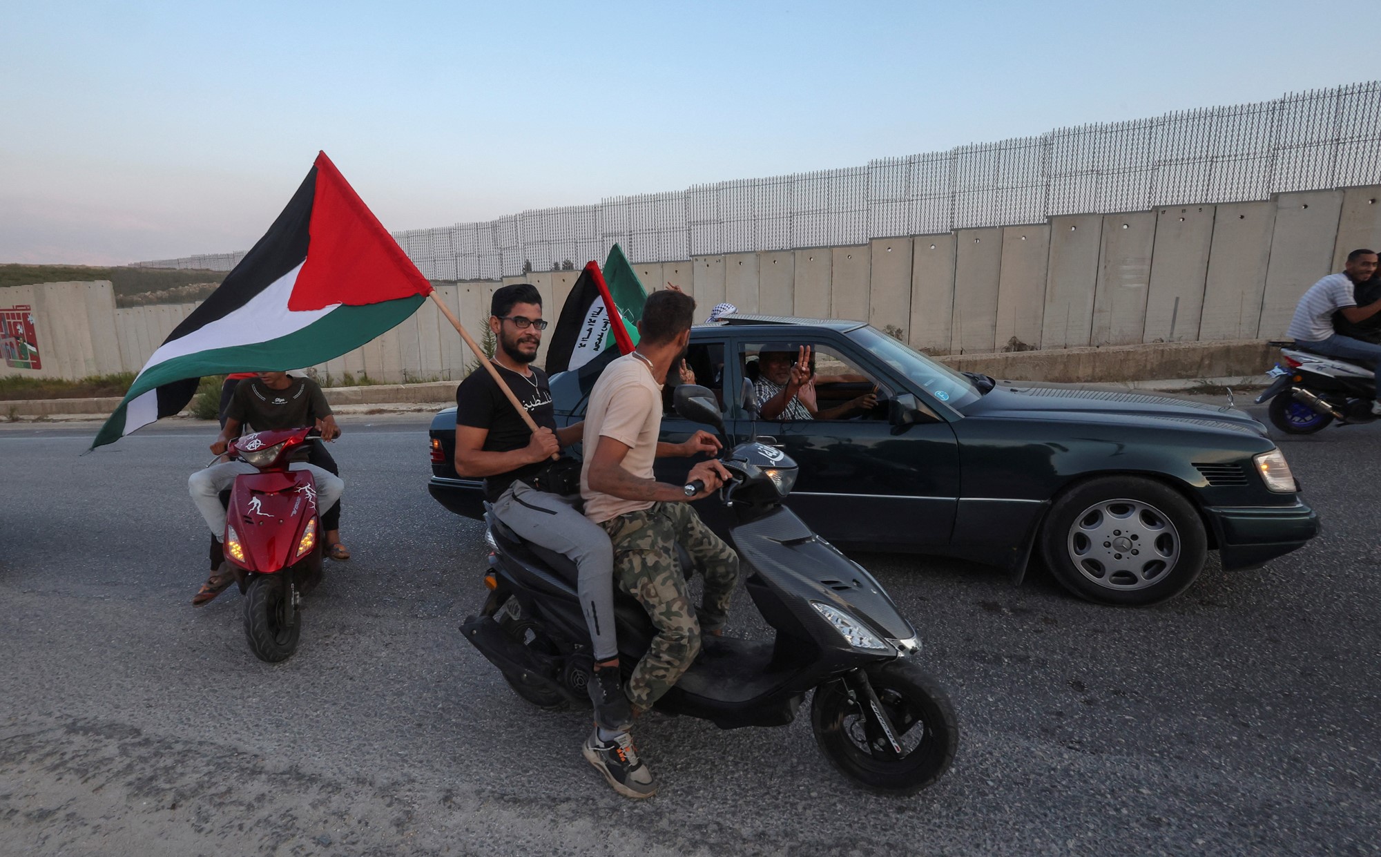 Two men on a motorbike, with one holding a flag, speak to men in a car.