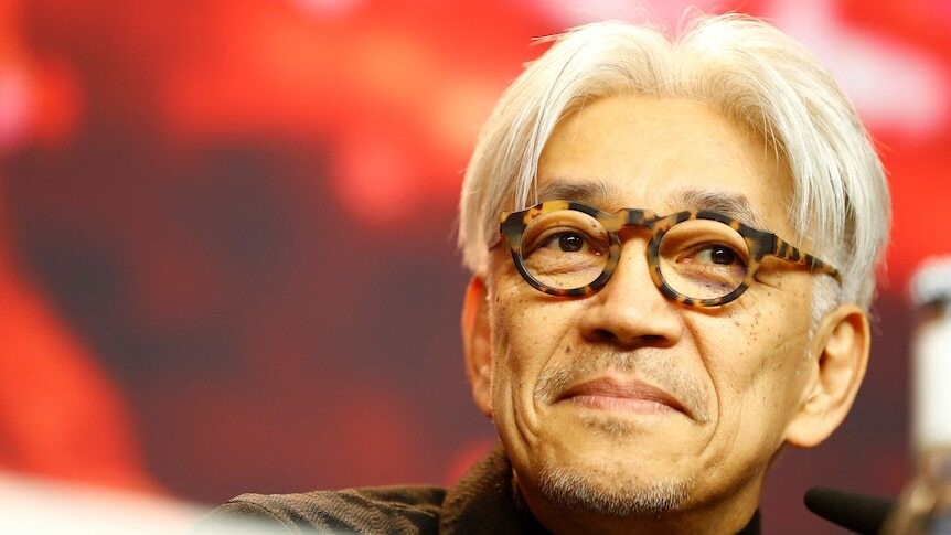 A close-up of an Asian man with white hair and round glasses, smiling without opening his mouth
