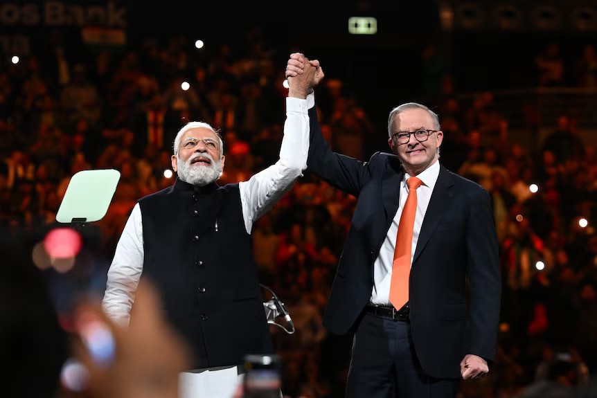 Narendra Modi and Anthony Albanese holding hands raised to the air, in an arena surrounded by a large crowd