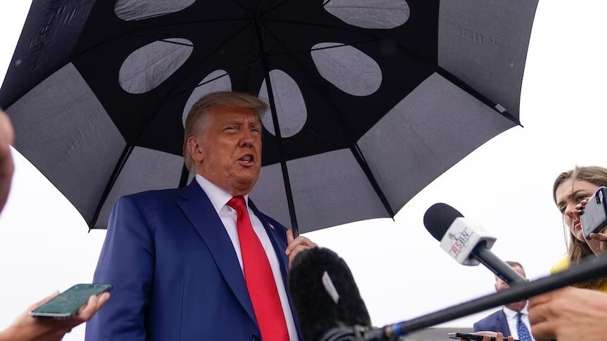 Donald Trump in a suit and tie, holding an umbrella and speaking with reporters