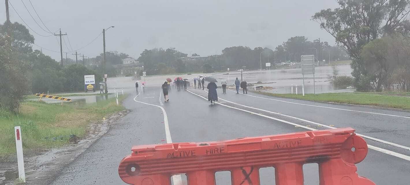 A dozen people walk past a barricade to look at a flooded road.