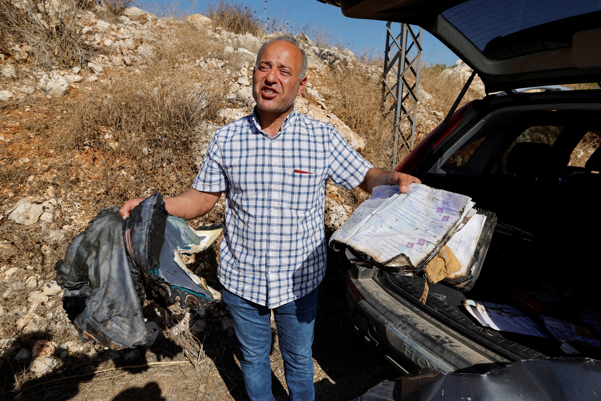 A man stands next to an open car boot holding what looks like a burnt backpack and a charred notebook or school book