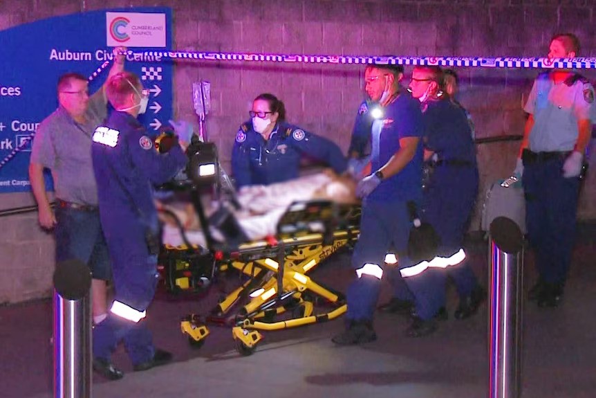 Emergency service workers pull a person on a stretcher out under police tape. The body on the stretcher has been blurred.