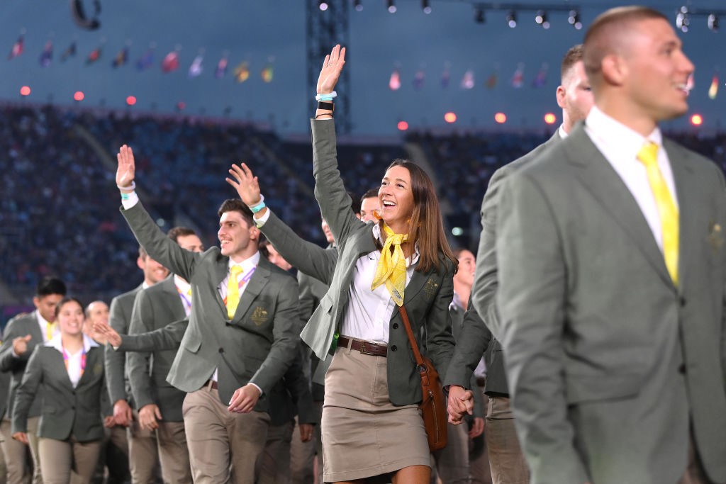 athletes from team australia walk in a row waving and smiling at the crowd during the opening ceremony