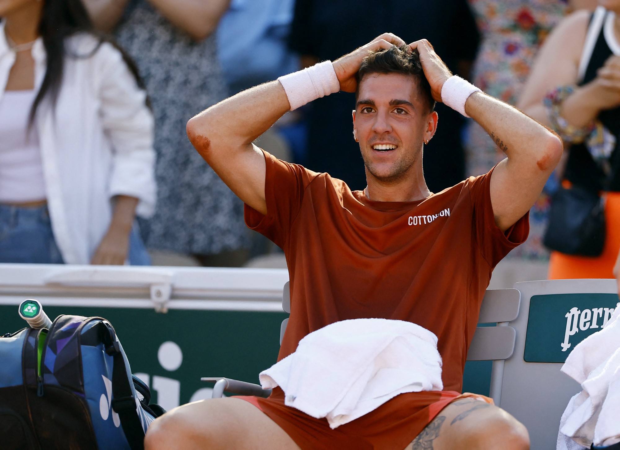 A male tennis player sits courtside after a game with his hands on his head and smiles