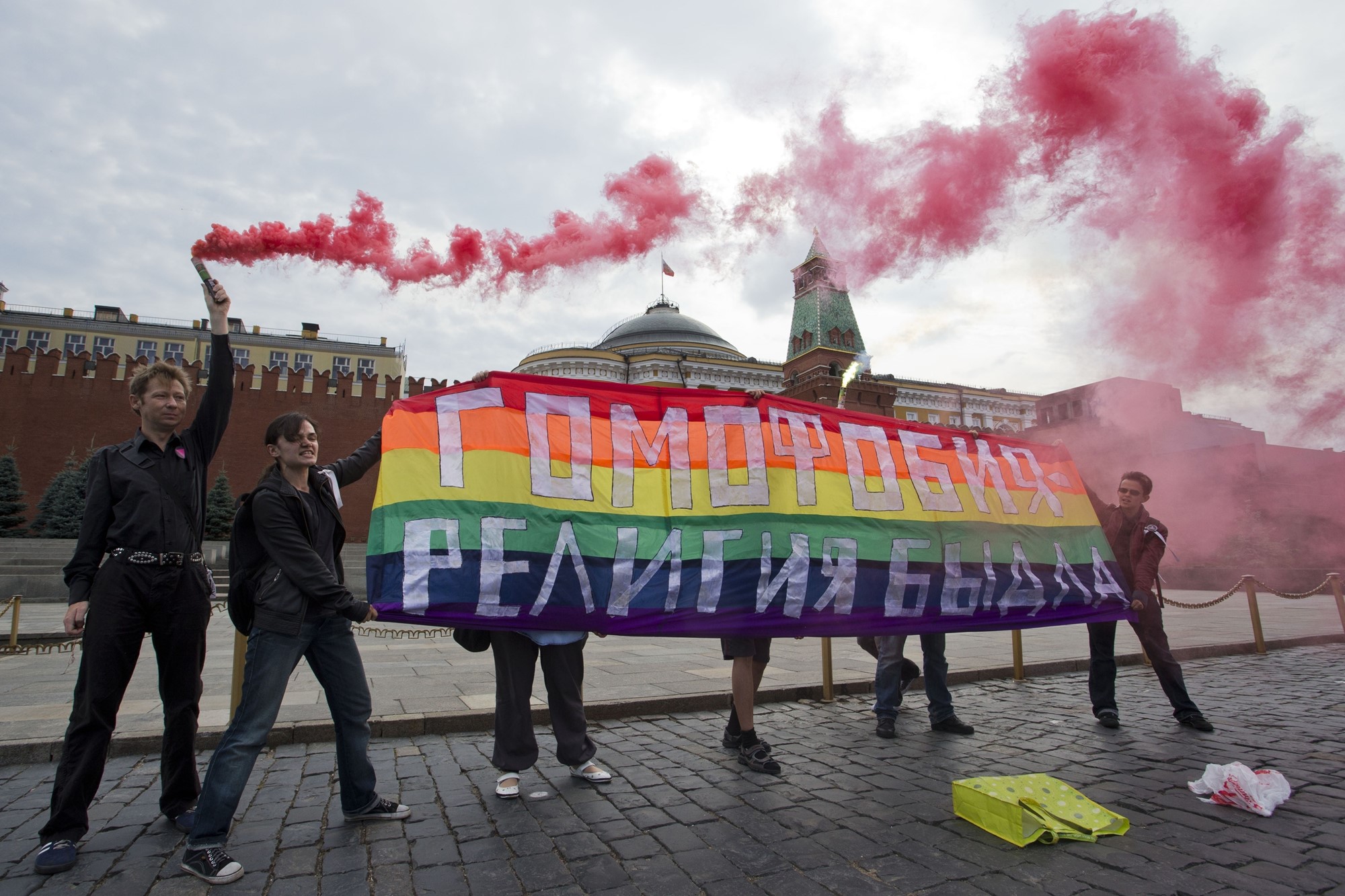 Multiple people hold a rainbow flag with Russian text on it. One person is letting off a flare with red smoke.