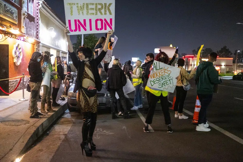 A group of strippers and supporters carry signs saying "Twerk Union" and "All workers deserve safety" outside a strip club.