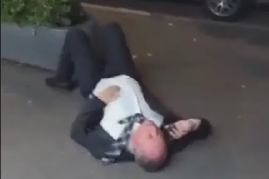 A screenshot of a man in a suit and tie lying on his back on a footpath and speaking on the phone