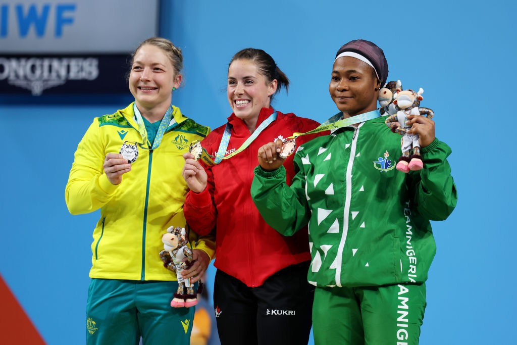 gold silver and bronze medalist weightlifters pose on podium with medals and mascots smiling