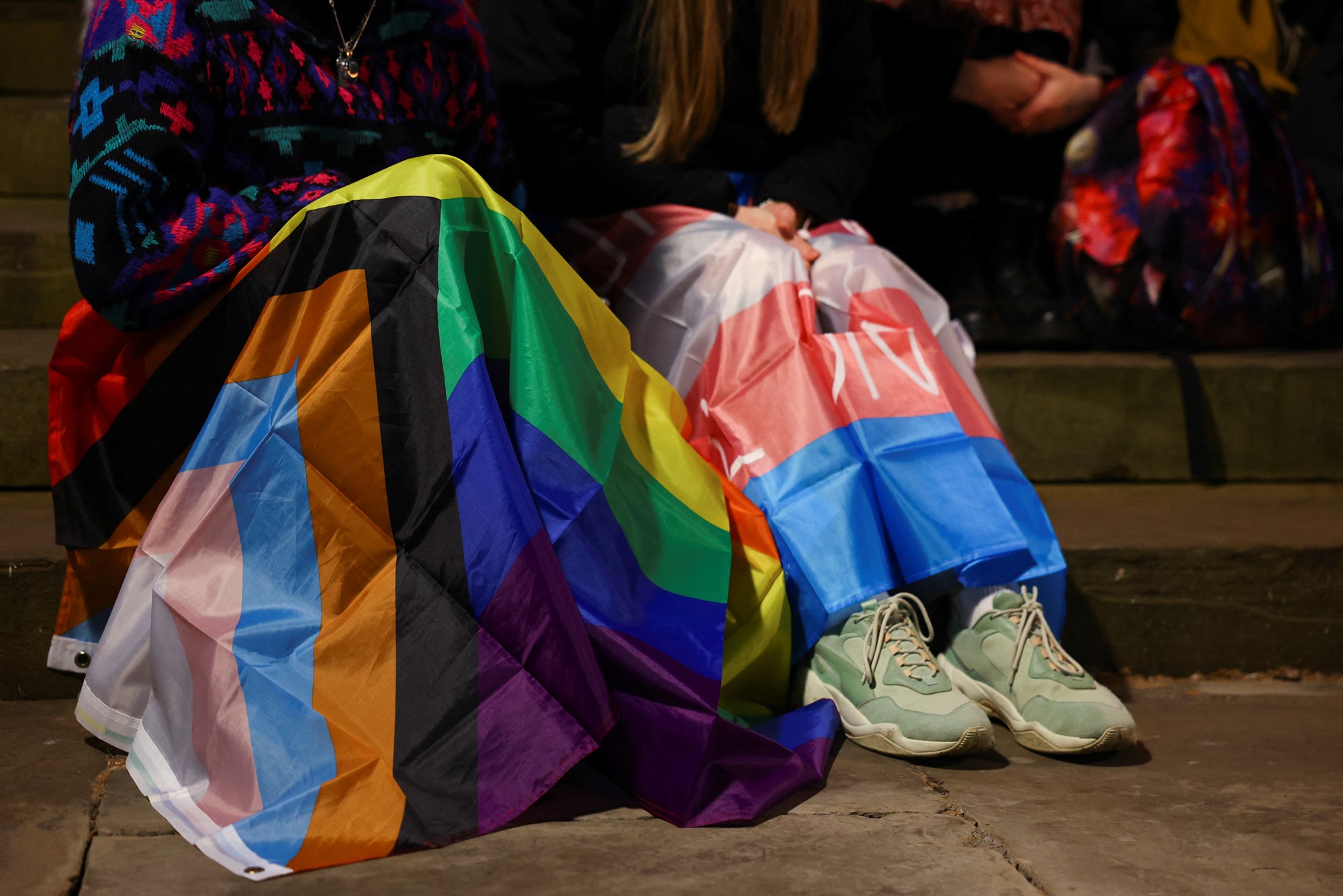 Two mourners sit together, one with the inclusive pride flag draped over their legs, the other with the trans flag.