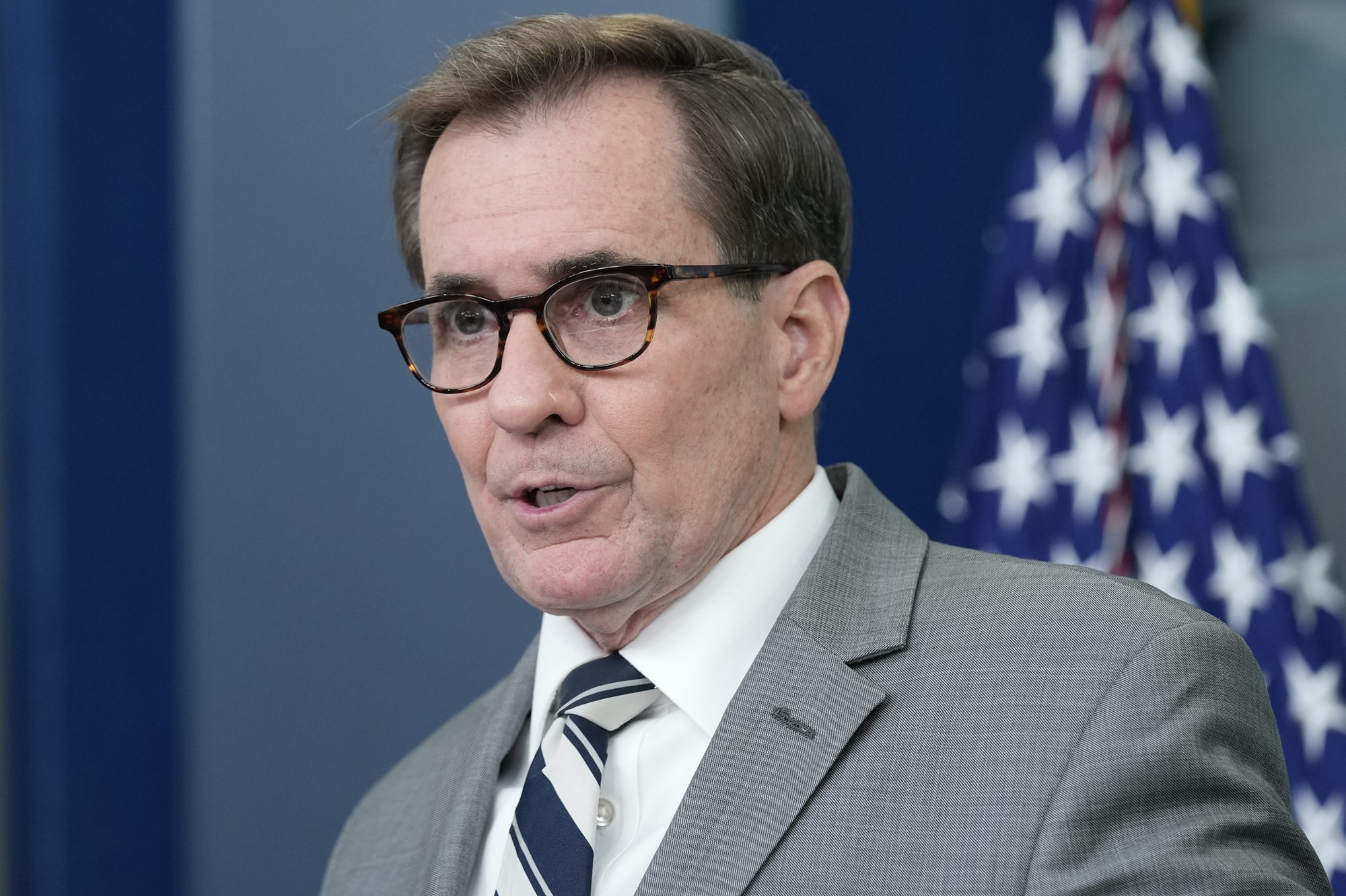 A middle aged white man in a suit and glasses speaks in front of an American flag