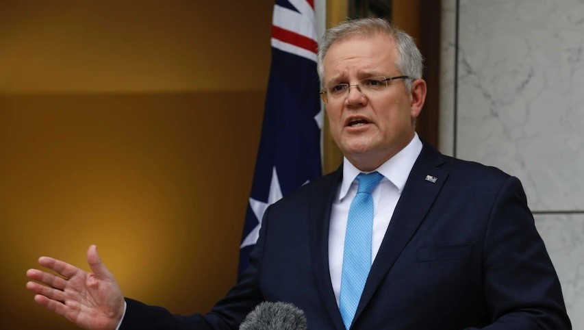 A man with grey hair and glasses in a suit and blue tie talks into a microphone.