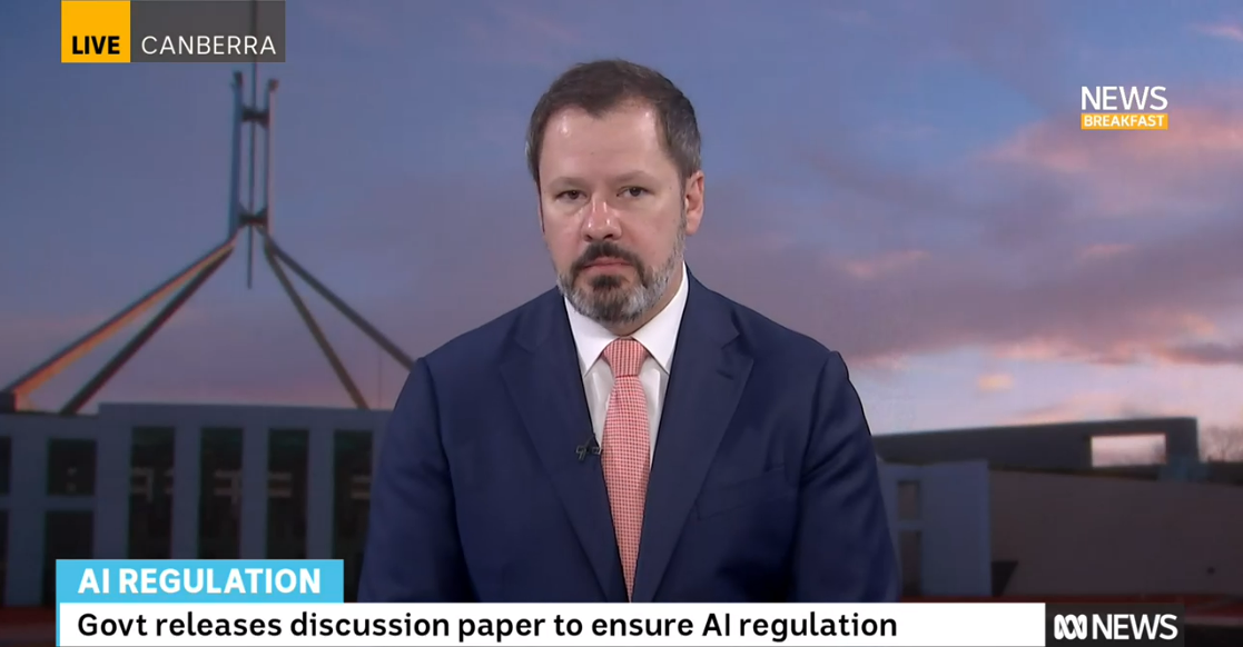 A middle-aged man in a suit does a TV interview in front of a backdrop showing parliament house in Canberra
