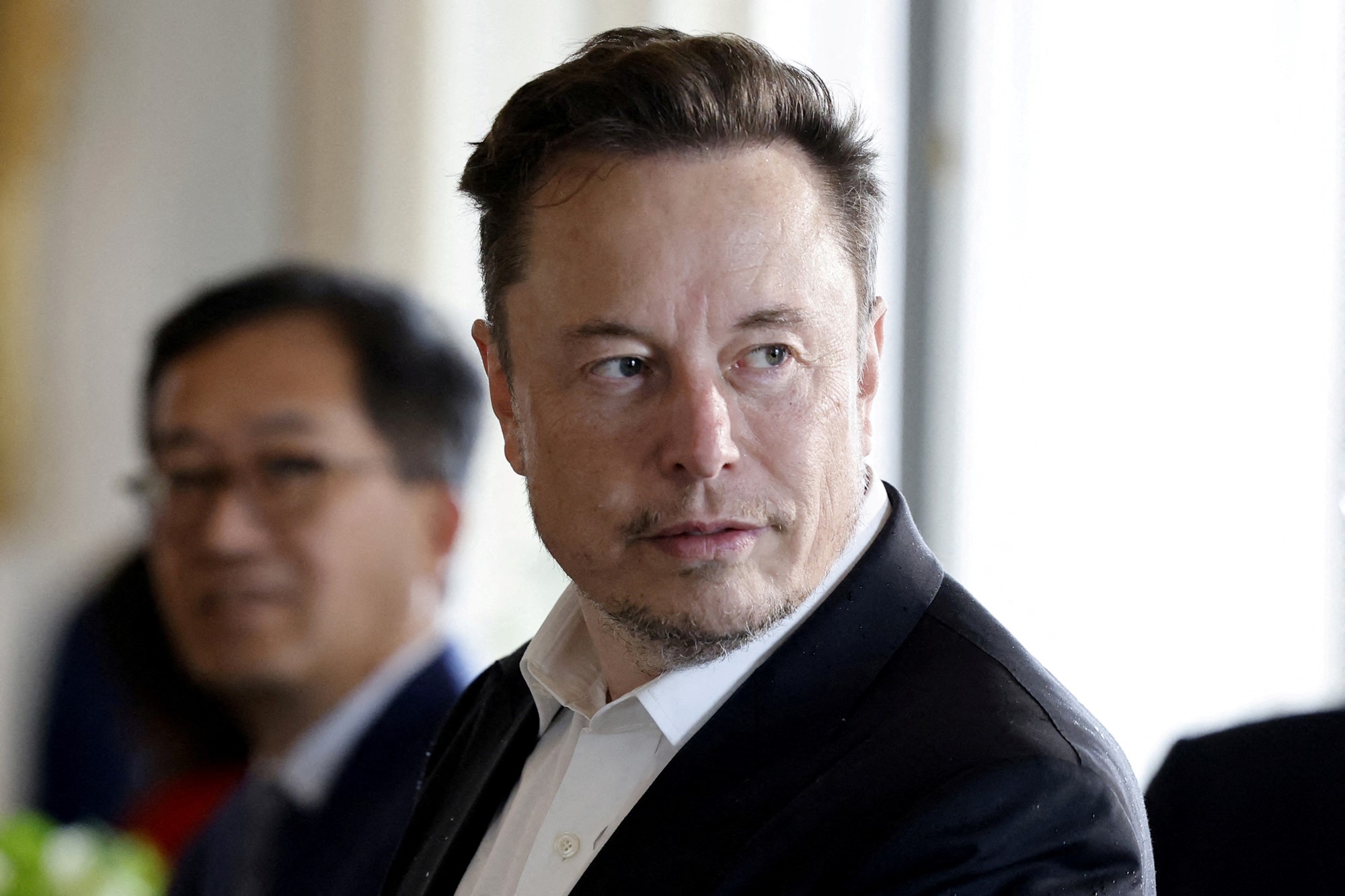 Elon Musk sits next to a man, both in suits