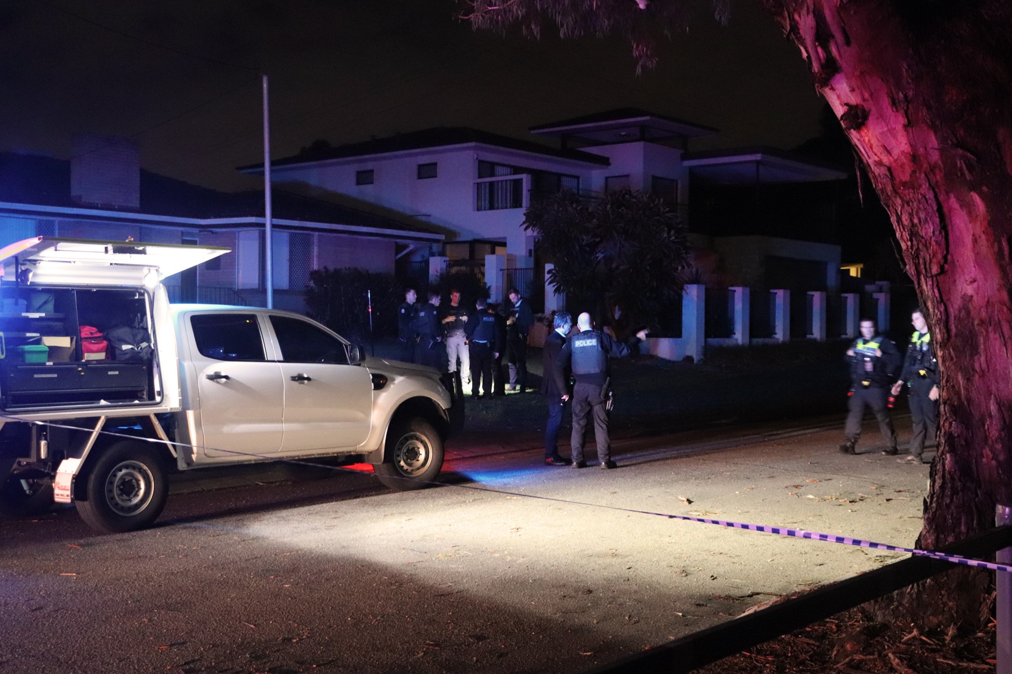 A white truck with a white light shines onto the road as police officers gather on the street