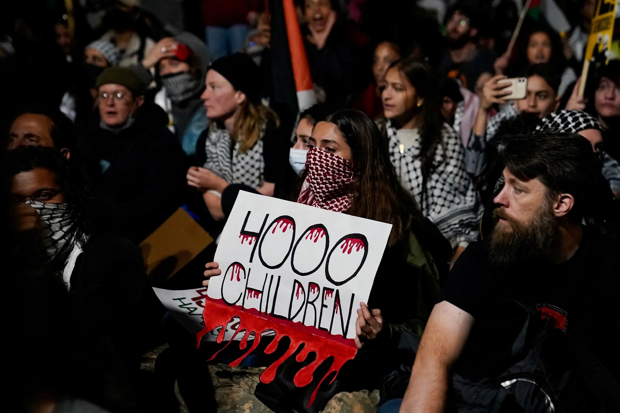 A female protester with her mouth covered with a Palestinian scarf holds a sign saying "4000 children" with dripping blood drawn on it