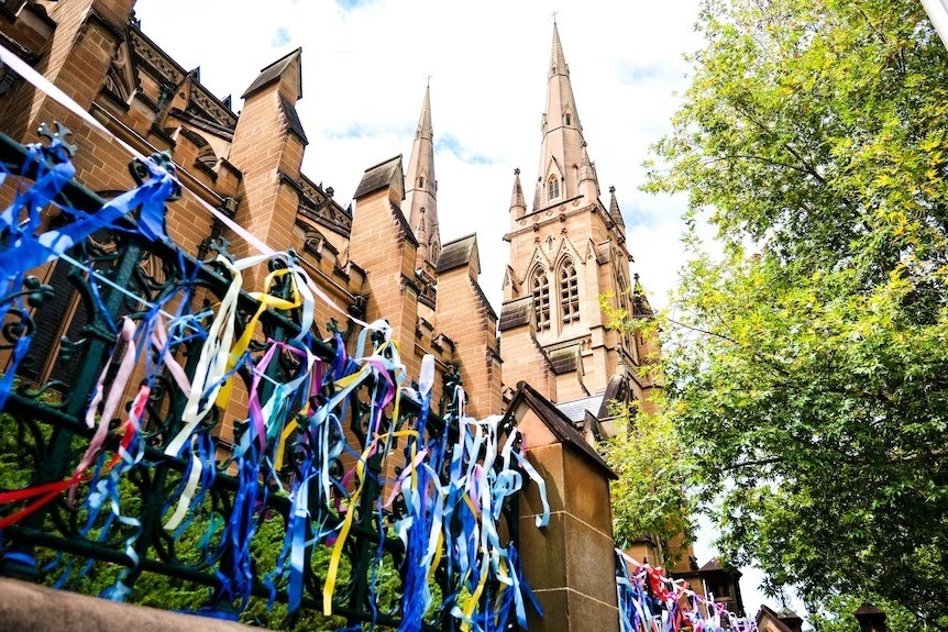 Hundreds of colourful ribbons tied to a wrought iron fence in front of a sandstone cathedral.