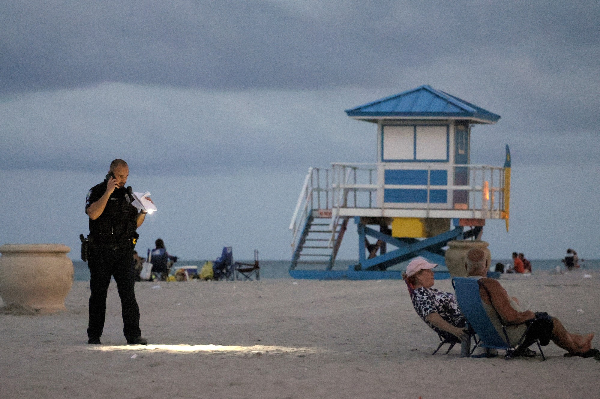 A police officer holding a torch on a beach in the evening, near beachgoers
