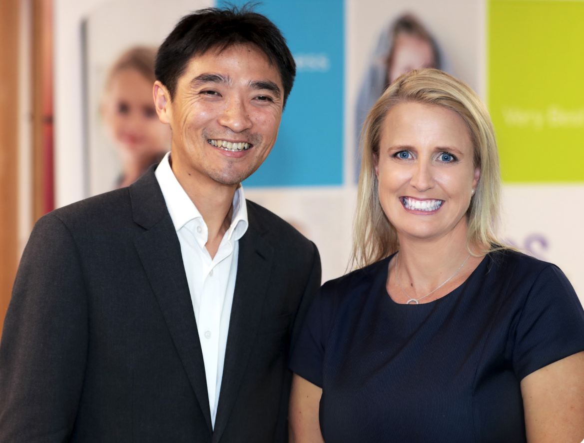 A man of Asian appearance in a black suit, next to a woman with blonde hair.