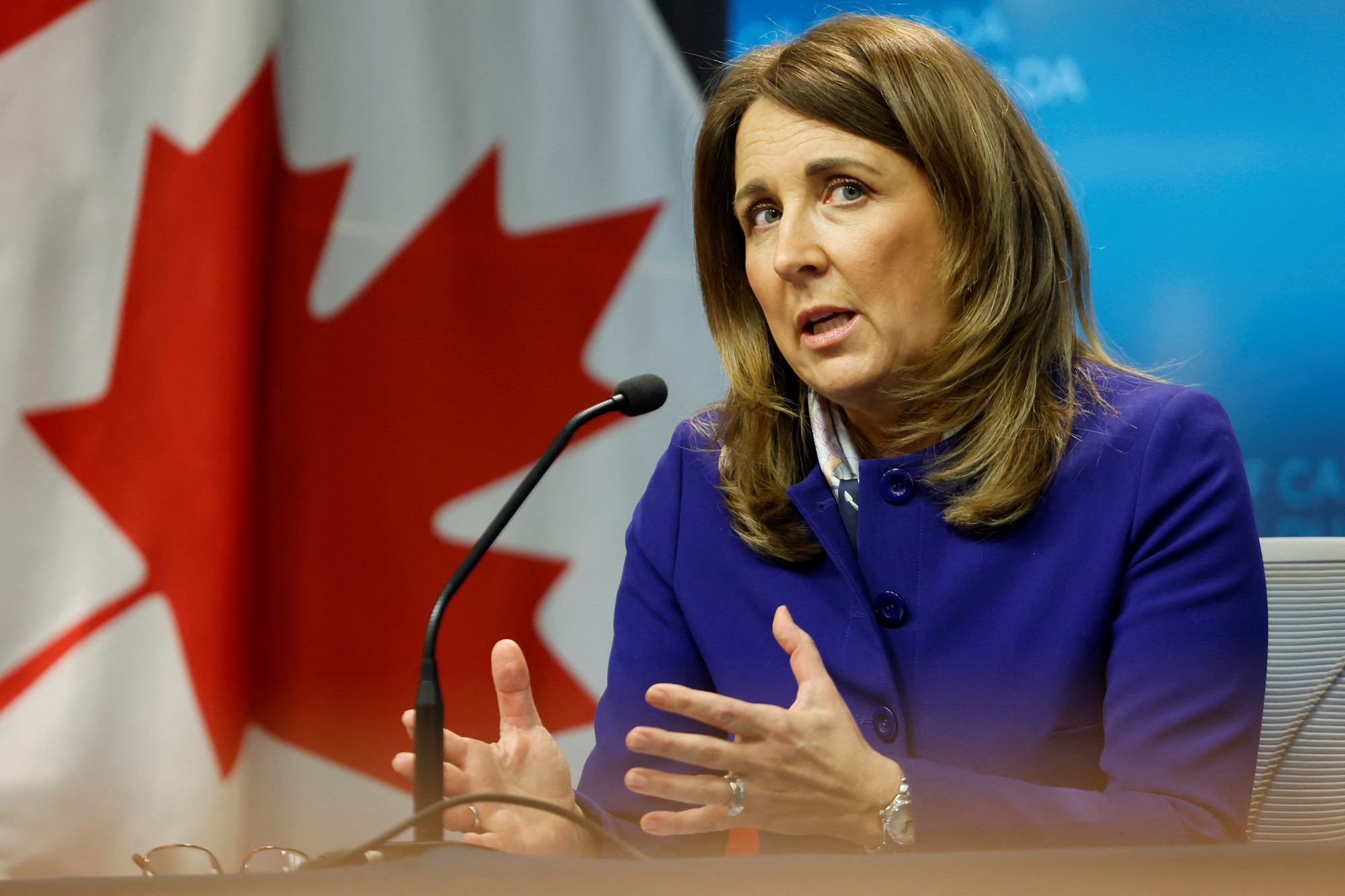 A woman wearing a blue jacket sits behind a small desk microphone talking and gesturing with her hands. A Canadian flag is in the background.