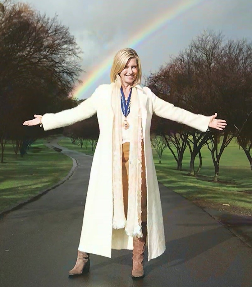 Olivia Newton-John smiles, standing beneath a rainbow.