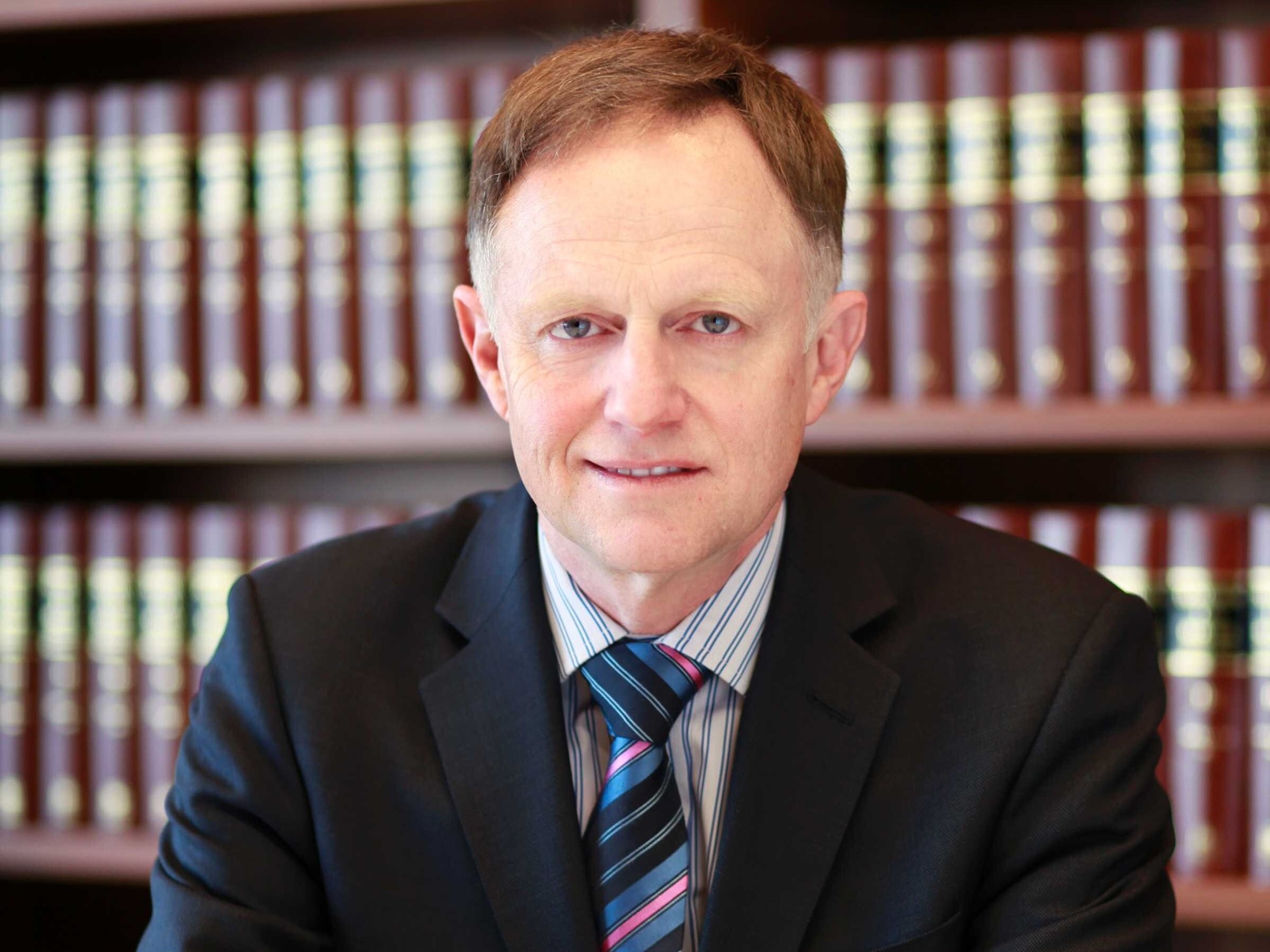 A man in a suit in front of a book shelf. 