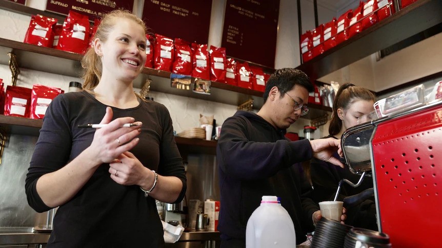 Two women and a man stand near a coffee machine, preparing coffees