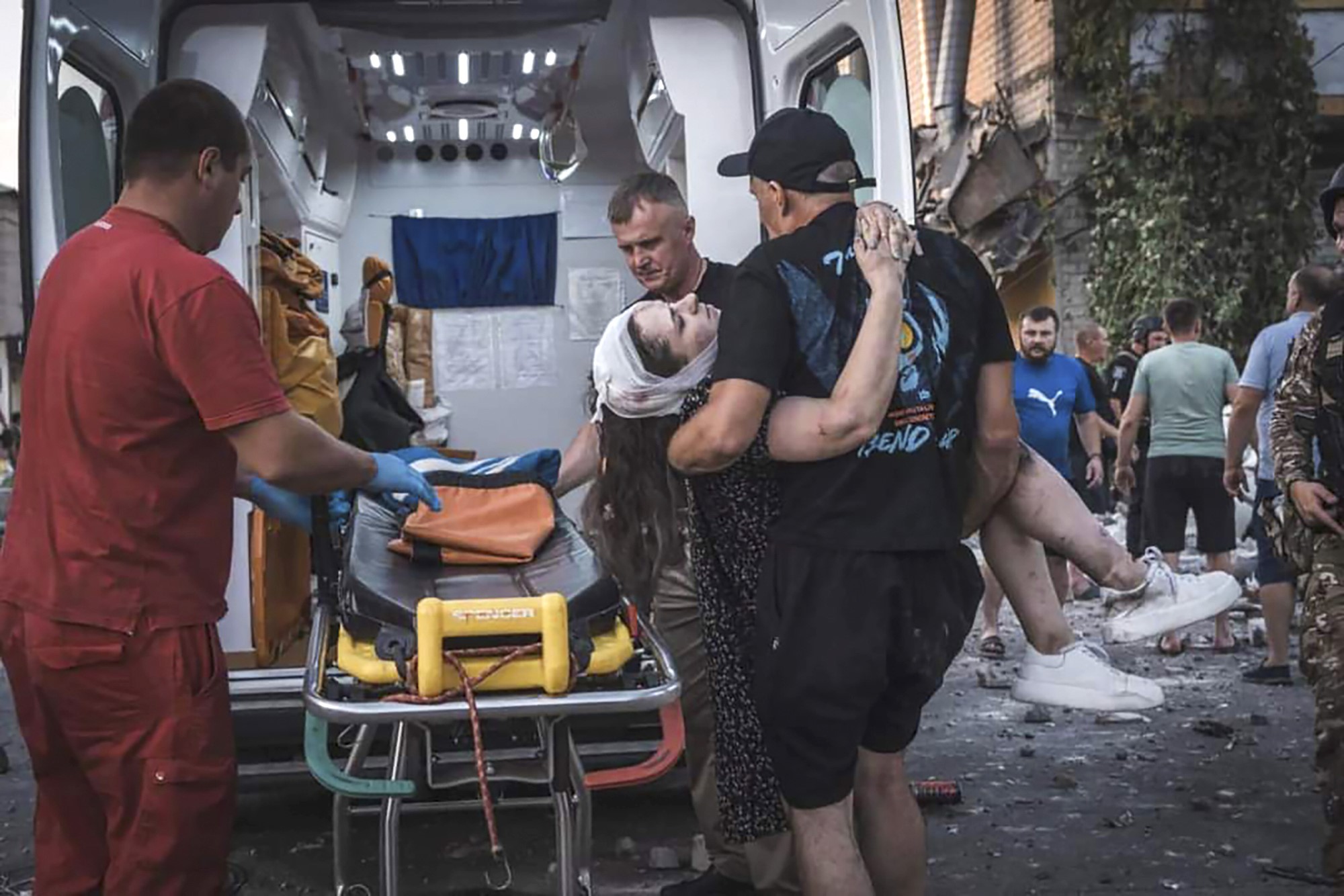 A man carrying a woman and placing her in a stretcher in a small ambulance, as emergency workers and volunteers work around them