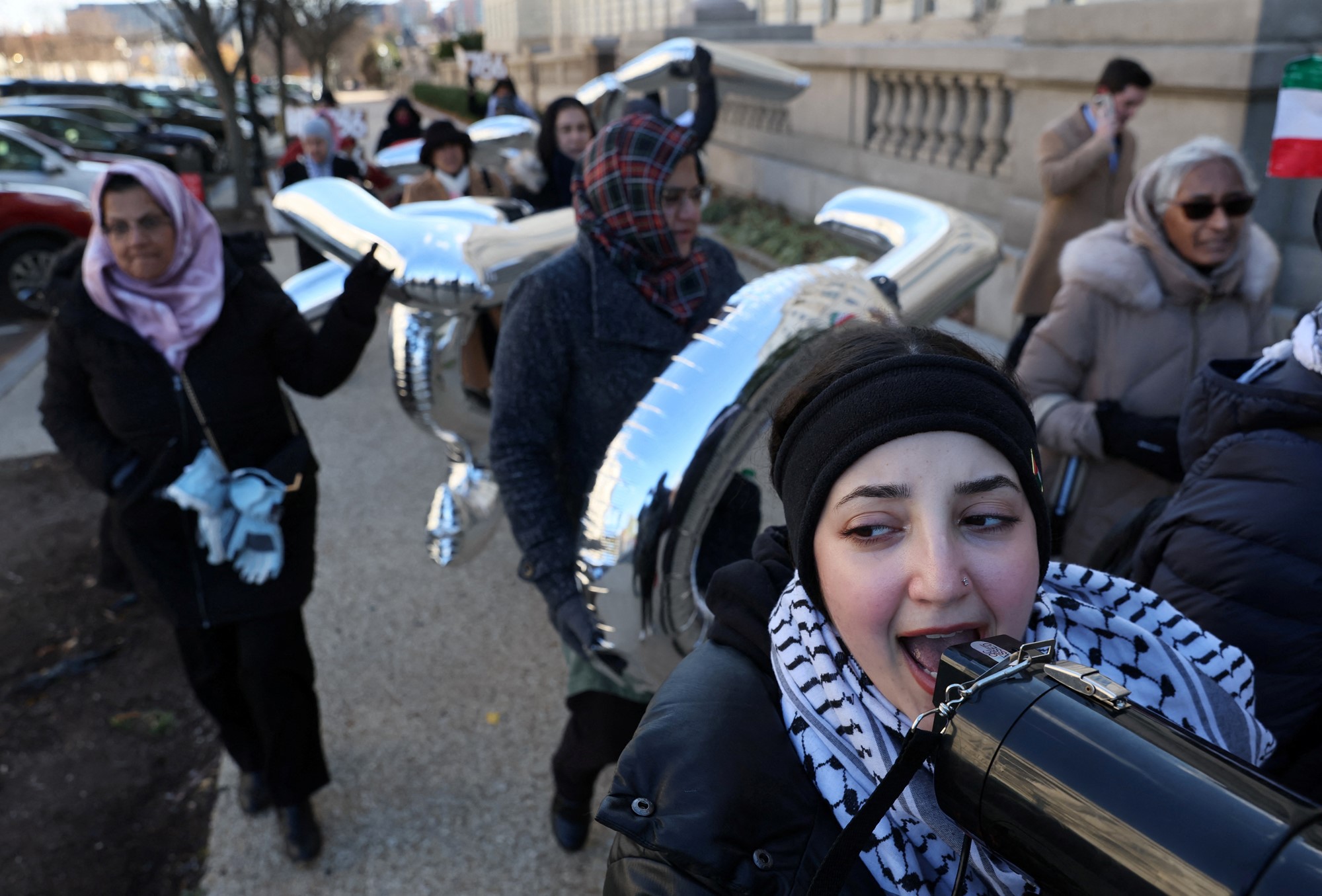 A close up of a group of women, one speaking into a megaphone, walking on a footpath with large inflatable letters and a Palestinian flag