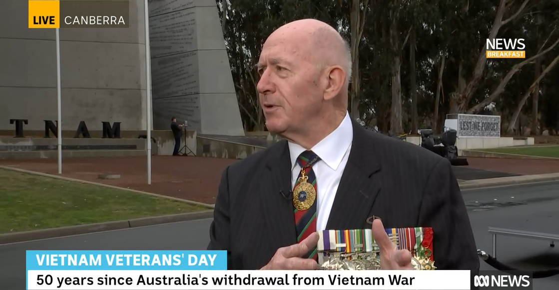 A bald man in a suit and tie wears army medals and gives a TV interview outside a war memorial