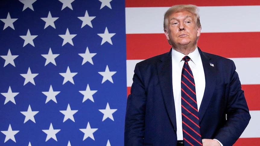 Donald Trump stands in front of an American flag. 