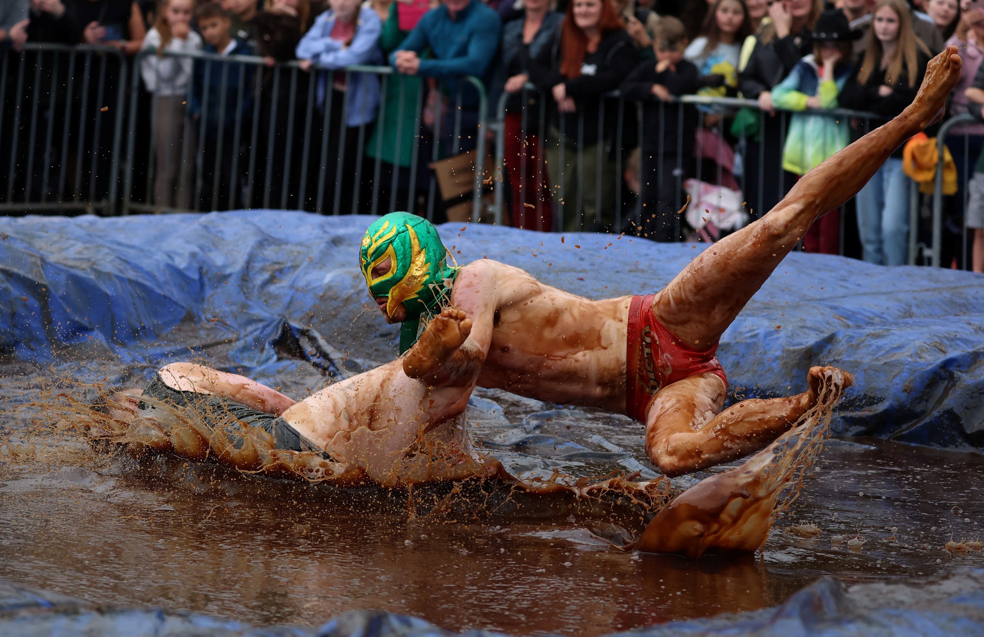 A man in a wrestling mask tackles another into a pool of gravy 